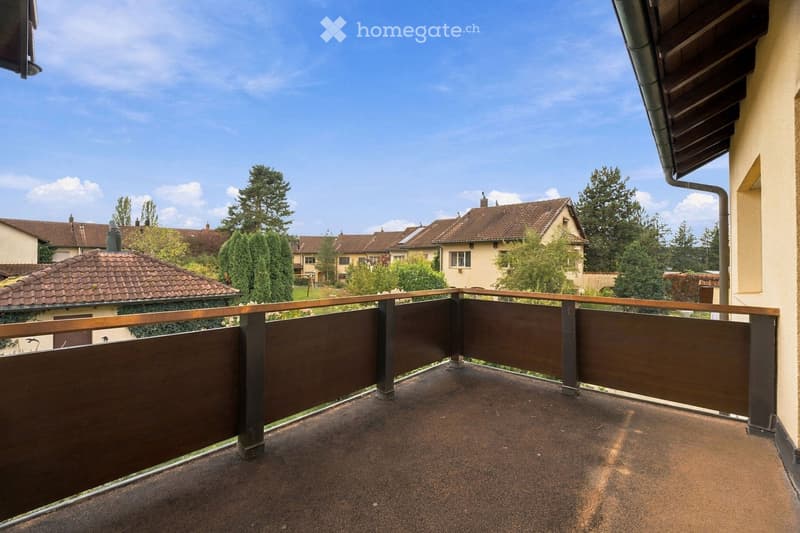 Balcony with wooden railing, overlooking a residential area with houses and trees under a clear blue sky.