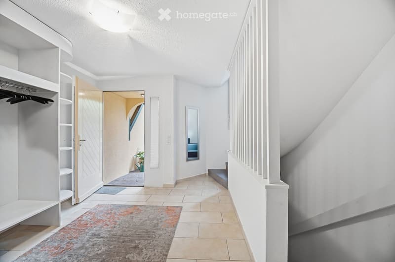 Modern hallway with white walls, tiled floor, built-in closet, staircase, and a small rug. A mirror and a plant are visible in the adjacent room.