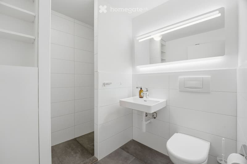 Modern, minimalist bathroom with white tiles, a white sink, a toilet, a large mirror with built-in lighting, and a sleek design.