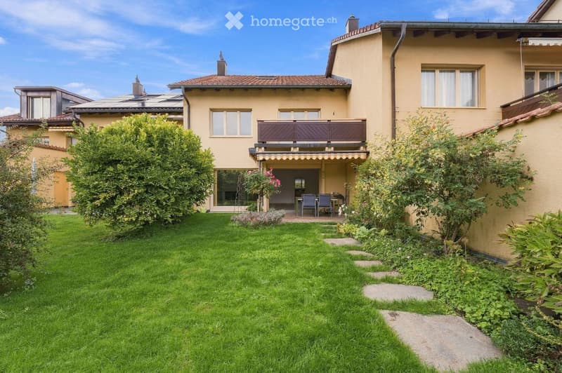 Well-maintained garden with green lawn, stone pathway, and small patio area with outdoor furniture near the house entrance.