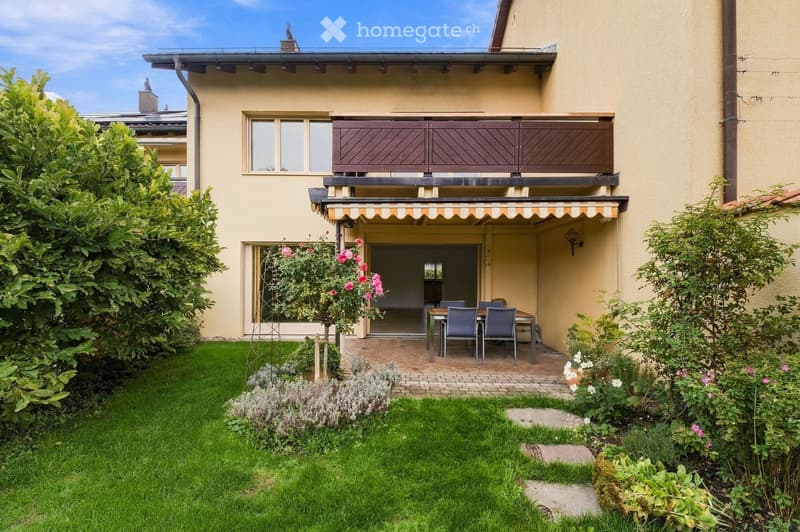 Garden area with a well-maintained lawn, flower beds, and a patio with a dining table and chairs. The house features a balcony with wooden shutters and a striped awning over the entrance.
