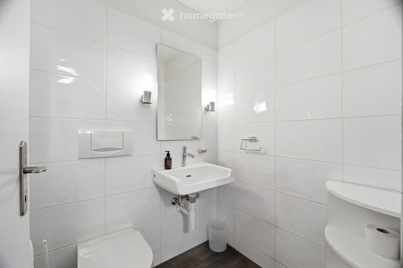 Modern bathroom with white tiles, a white sink with a chrome faucet, a mirror above the sink, a toilet, a towel rack, and a soap dispenser.