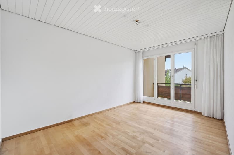 Empty room with white walls, wooden flooring, large window with white curtains, and a sliding glass door leading to a balcony.
