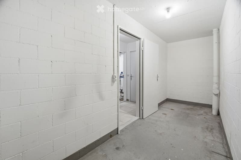 A narrow, white-walled corridor with a concrete floor, leading to an open doorway that reveals a glimpse of another room. The walls are made of white bricks, and the ceiling is white with a single light fixture.