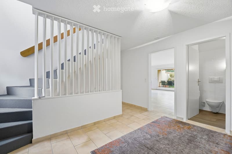 Modern hallway with white walls, tiled floor, staircase with white balustrade, and an open doorway leading to a bathroom and outdoor area.