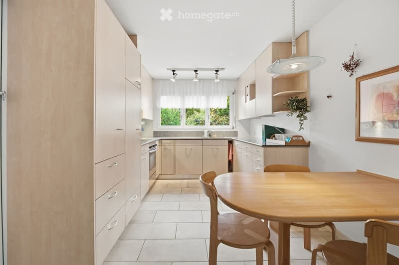 Bright kitchen with light wood cabinets, a round wooden dining table with chairs, and a pendant light hanging above the table. The kitchen features a white tiled floor, a window with white curtains, and a small plant on a shelf.