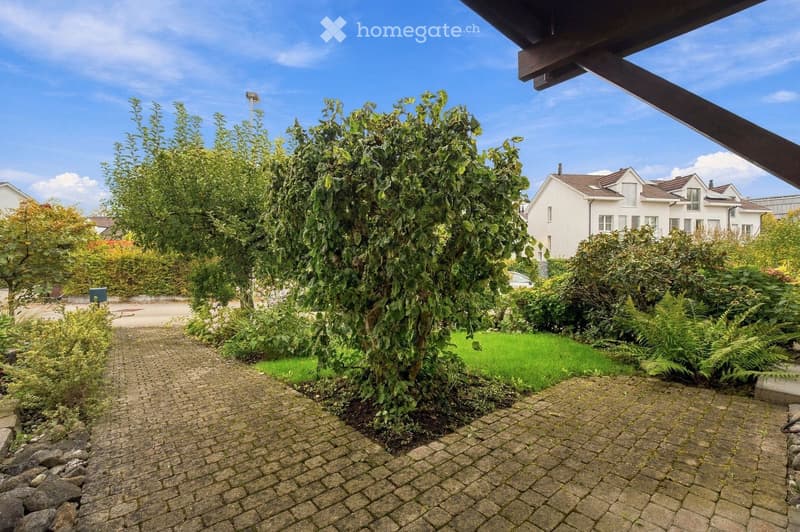 Garden area with a brick pathway, lush greenery, and a large house in the background.