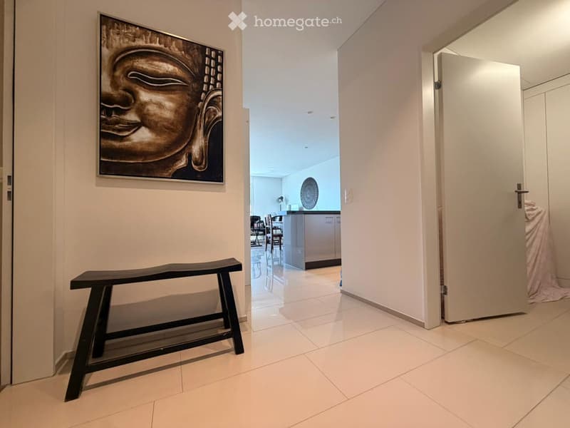 A modern hallway with white tiled flooring, a black bench, and a large Buddha head artwork on the wall. The hallway leads to a kitchen area with white cabinets and a dining table with chairs.