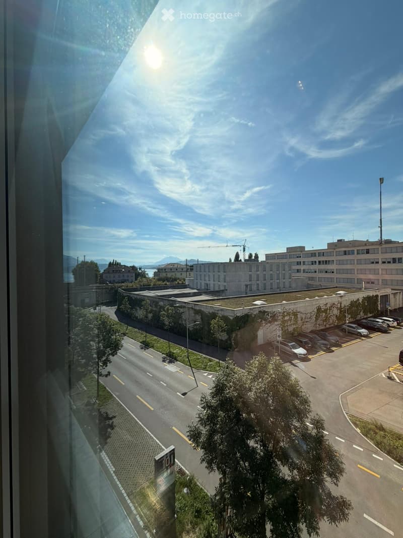 View from a high-rise window showing a multi-story building, parking lot, road, and distant landscape with mountains and water.