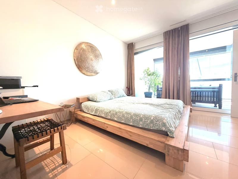 Bright bedroom with a wooden bed, patterned bedding, a wooden table with a laptop, a woven chair, and a decorative wall piece. Large windows with curtains lead to a balcony.