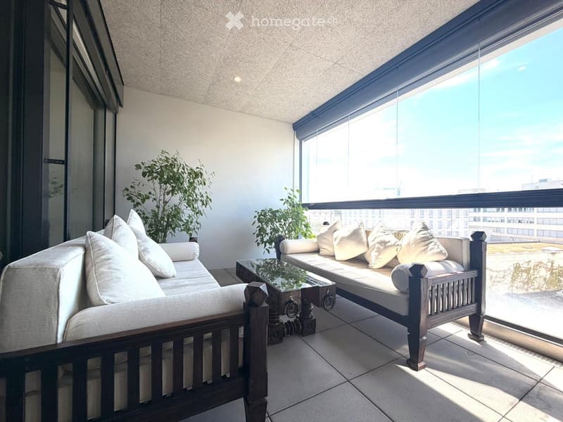 Modern balcony with large windows offering a city view, featuring a sofa, coffee table, and potted plants.