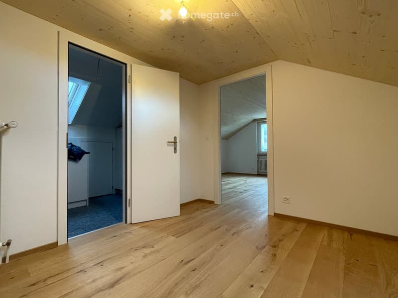 Empty room with wooden flooring, white walls, and two open doors leading to other spaces. The room has a slanted wooden ceiling and visible electrical outlets.