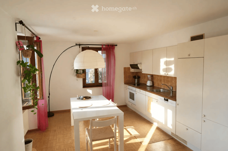 Modern kitchen with white cabinets, wooden floor, white dining table with chairs, and a hanging lamp. Features a sink, oven, dishwasher, and a blender on the counter.