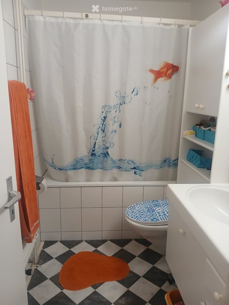 Bathroom with white tiles, a bathtub with a decorative shower curtain featuring a goldfish and water waves, a toilet with a blue patterned seat cover, a sink with a cabinet, and an orange towel on the floor.