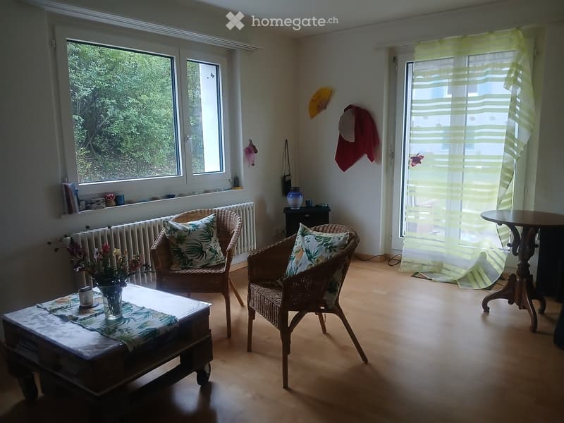 Bright living room with wooden flooring, two wicker chairs with floral cushions, a coffee table with a vase of flowers, and a small round table near the window.