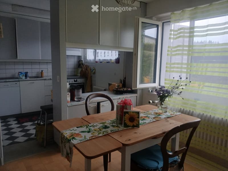 Bright kitchen and dining area with wooden table, chairs, and a vase of flowers. The kitchen features white cabinets, a dishwasher, and an oven.