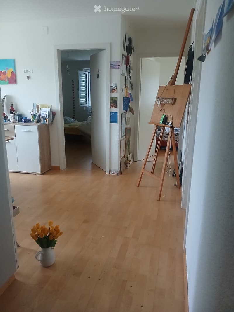 Bright hallway with wooden flooring, leading to adjacent rooms. Features a vase with yellow tulips, an easel, and a cabinet with various items.