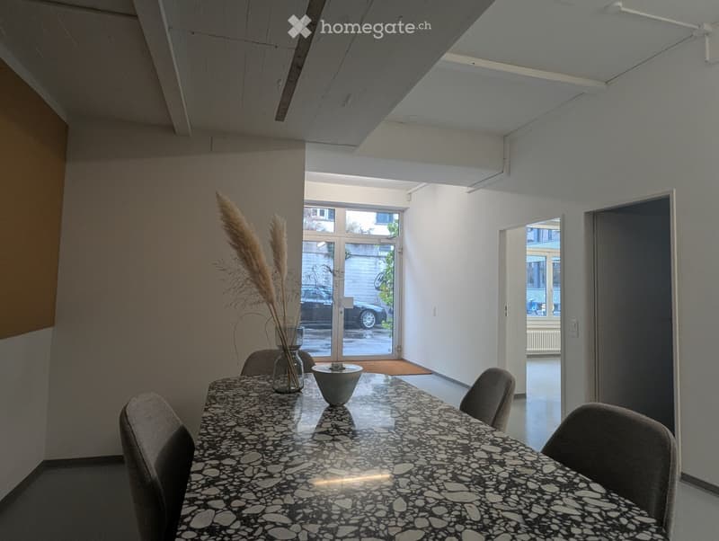 Bright, minimalist room with a long black and white speckled table surrounded by four grey chairs, a vase with tall dried plants, and a decorative bowl on the table. Large windows and doors lead to an outdoor area.