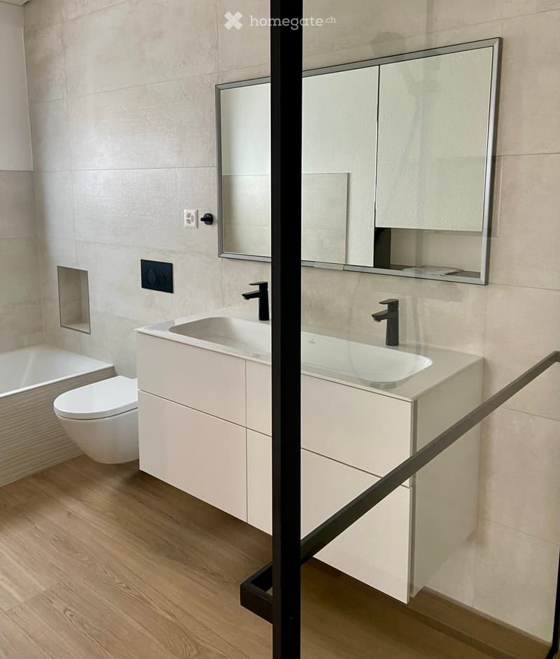 Modern bathroom with a double sink vanity, sleek faucets, a large mirror, a toilet, and a bathtub, all set against light-colored tiles and wooden flooring.