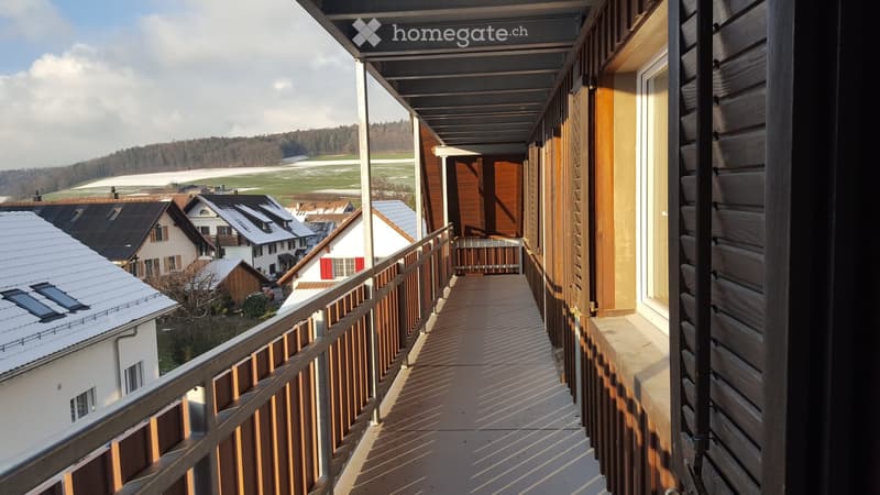 Wooden balcony with railing overlooking a rural landscape with houses and fields.