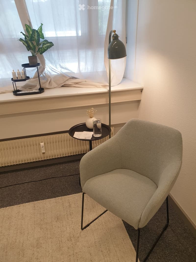 A modern, minimalist room with a grey armchair, small black side table, floor lamp, and a potted plant near the window.