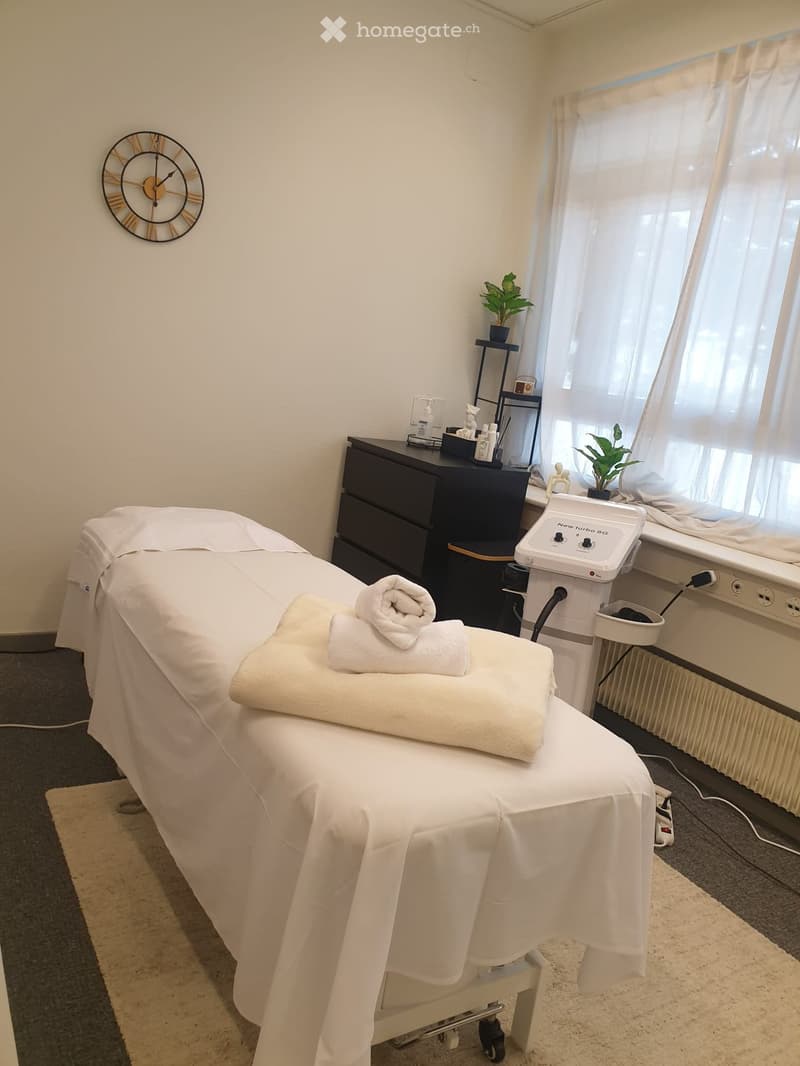 A well-lit massage room with a massage table covered in white linens, neatly folded towels, a small cabinet, a potted plant, a wall clock, and a massage machine on a stand.