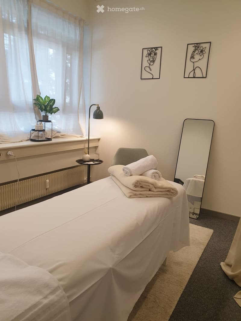 A neatly arranged bedroom with a white bed, neatly folded towels, a side table with a lamp, a plant on the windowsill, and two decorative picture frames on the wall.