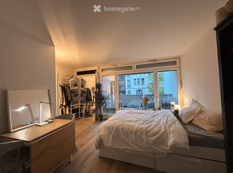 Cozy bedroom with a large bed, wooden floor, large window with a view, and a small desk with a mirror and lamp.