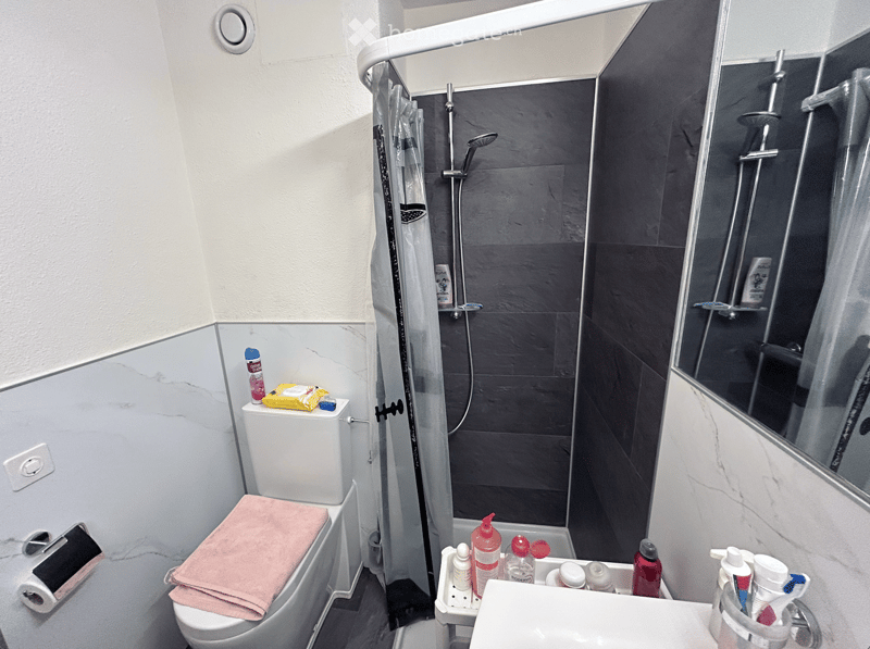 Modern bathroom with a toilet, sink, and shower area. Features a marble-patterned wall, a glass shower enclosure, and a towel on the toilet seat.