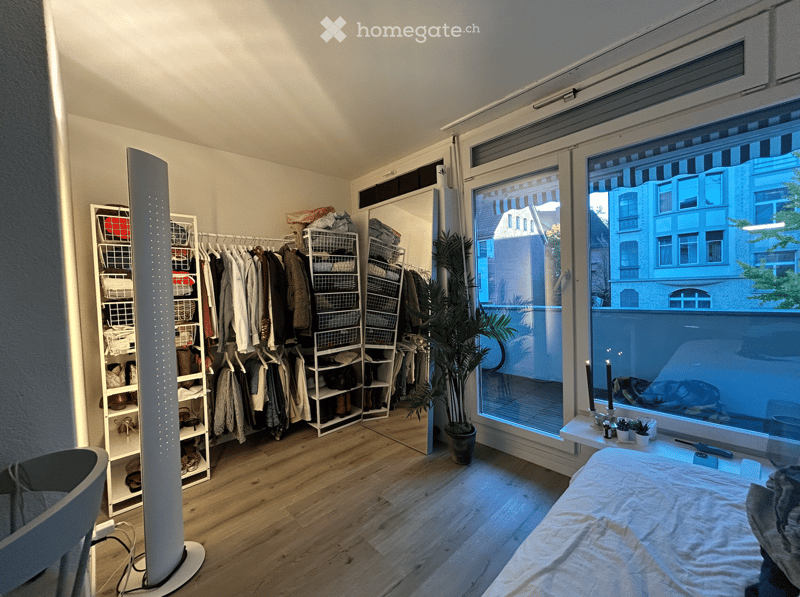 Well-lit bedroom with wooden flooring, large window offering city view, bed with white linens, wardrobe with hanging clothes, shoe rack, potted plant, and decorative candles on windowsill.