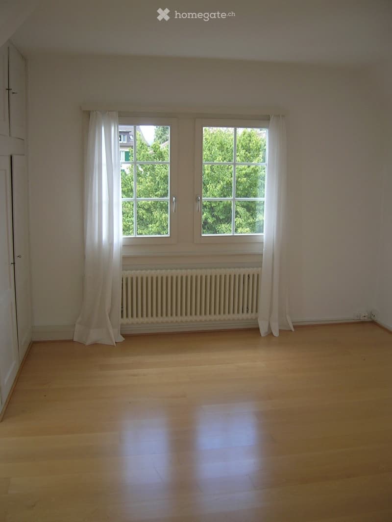 Empty room with wooden flooring, white walls, two windows with white curtains, and a radiator below the windows.