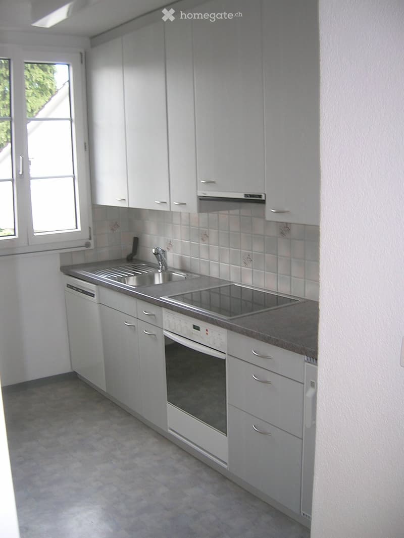 Modern kitchen with white cabinets, stainless steel sink, oven, and dishwasher. Light-colored tiles and a window provide natural light.