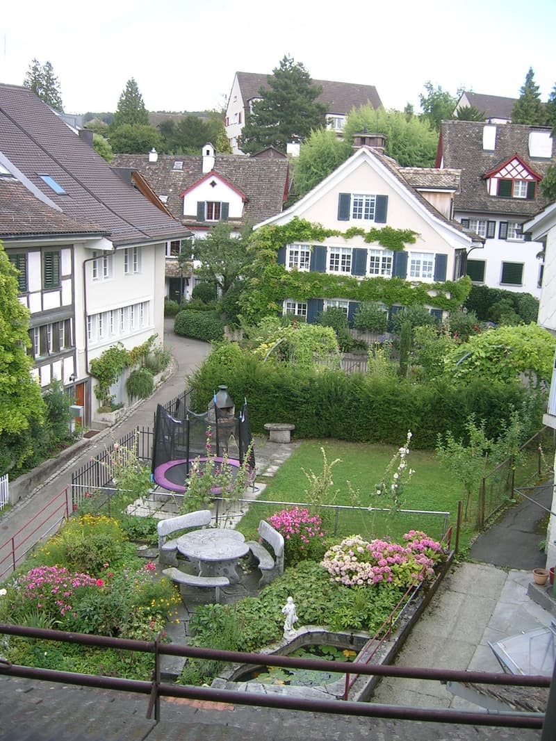 A lush garden with a round table, benches, flowers, a small pond, and a trampoline, surrounded by residential houses.