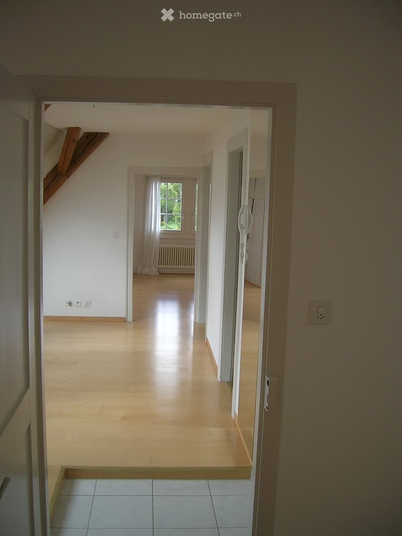 Empty hallway with light wooden flooring, white walls, and a staircase visible on the left side. A window with white curtains is seen at the end of the hallway.