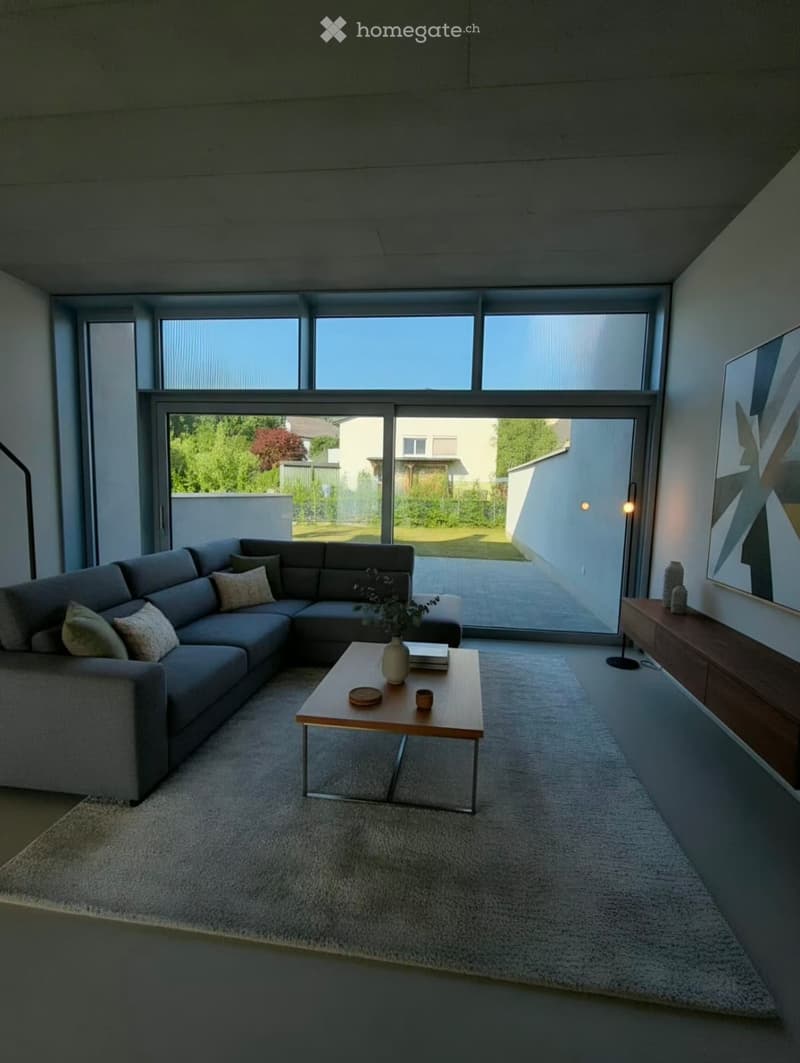 Modern living room with large windows and sliding glass doors leading to an outdoor area. Features a grey sectional sofa, wooden coffee table, floor lamp, sideboard, and decorative items.