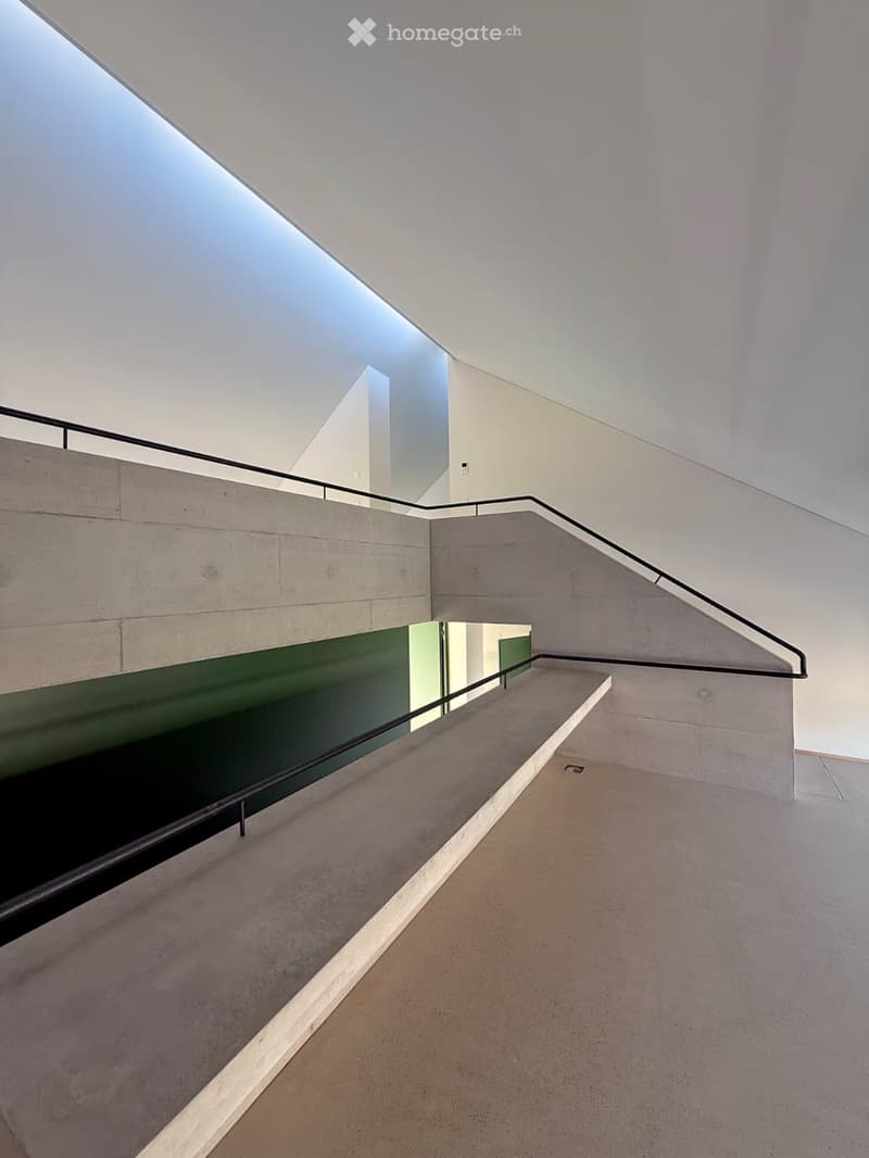 Modern, minimalist hallway with concrete walls, a staircase, and a green accent wall. The ceiling features a skylight, allowing natural light to illuminate the space.