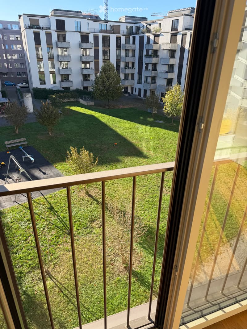 Balcony overlooking a green courtyard with trees, benches, and playground equipment. Modern apartment buildings surround the area.