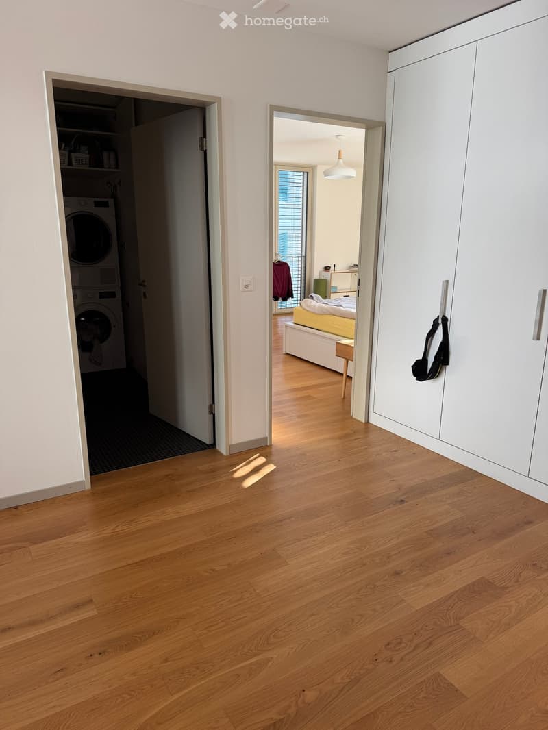 Empty hallway with light wooden flooring, white walls, and open doors leading to a bedroom and a laundry area.