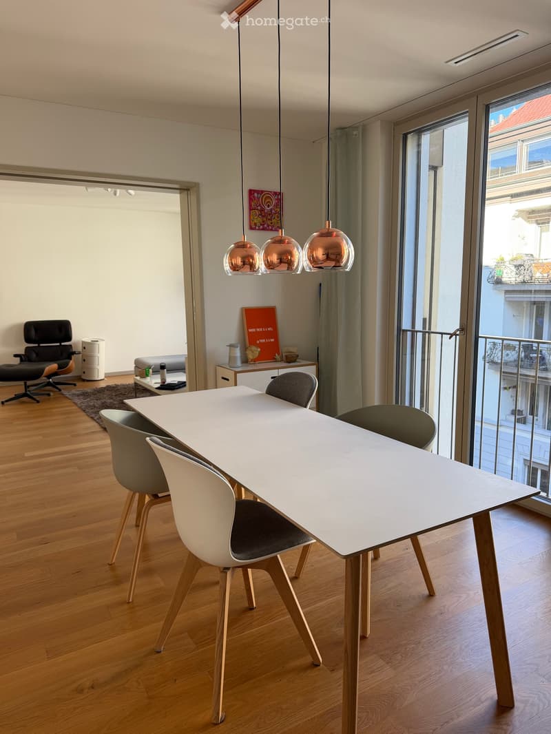 Modern dining area with a white dining table, four chairs, pendant lights, a sofa, and a cabinet with decorative items.