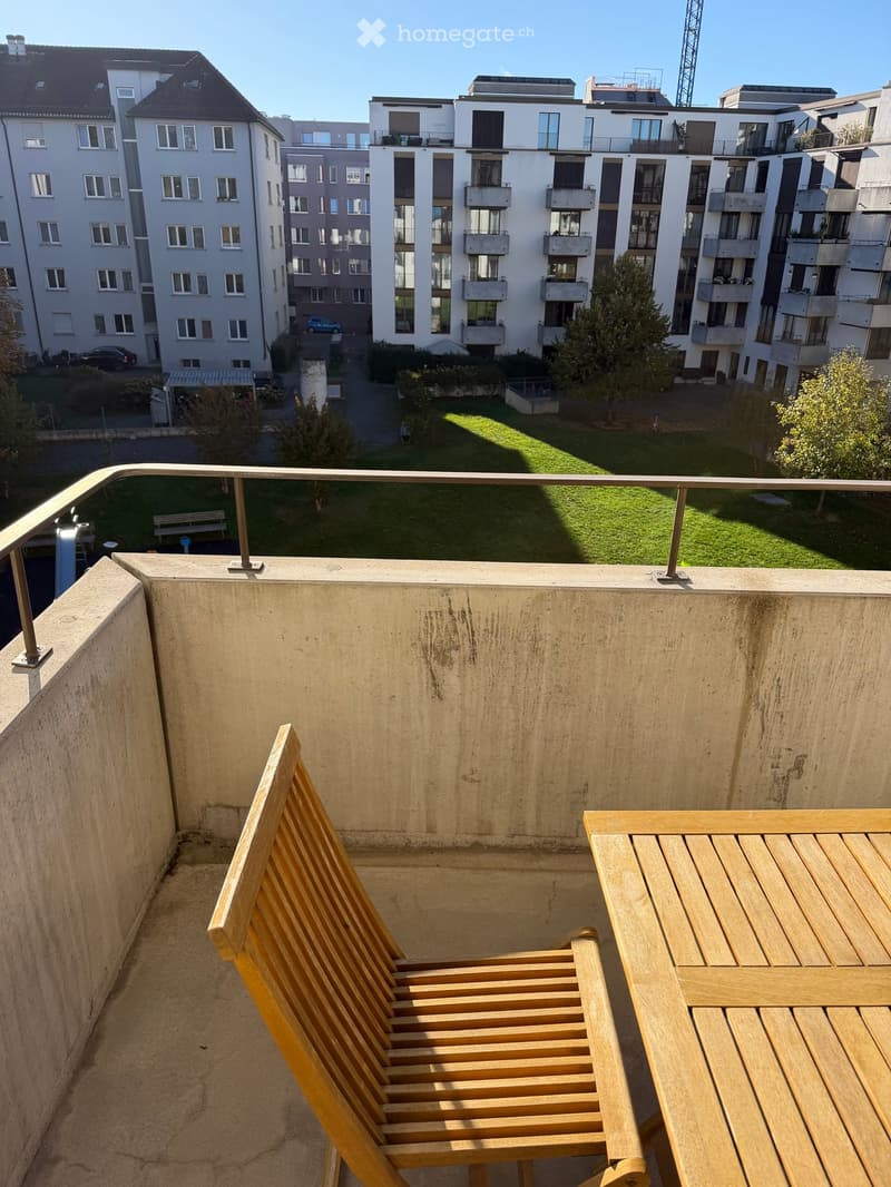 Balcony with wooden chair and table, overlooking a green lawn and modern apartment buildings.