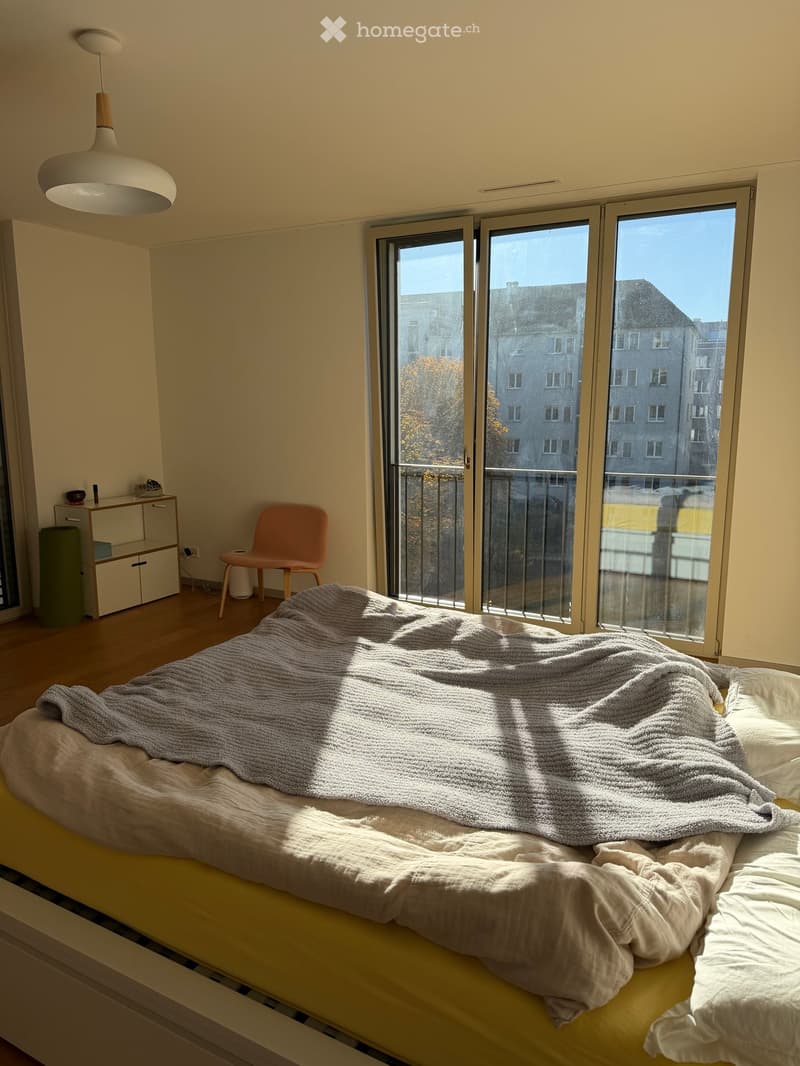 Bright bedroom with large windows offering a view of the city, featuring a bed with a blanket, a chair, a small cabinet, and a hanging lamp.