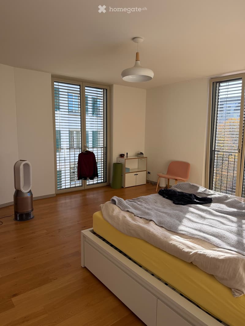 Modern bedroom with wooden flooring, large windows with blinds, a bed with a grey blanket, a chair, a fan, and a small cabinet.