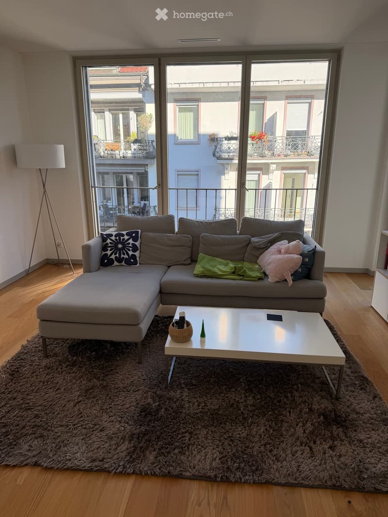 Modern living room with a grey sectional sofa, white coffee table, floor lamp, and large windows opening to a balcony.