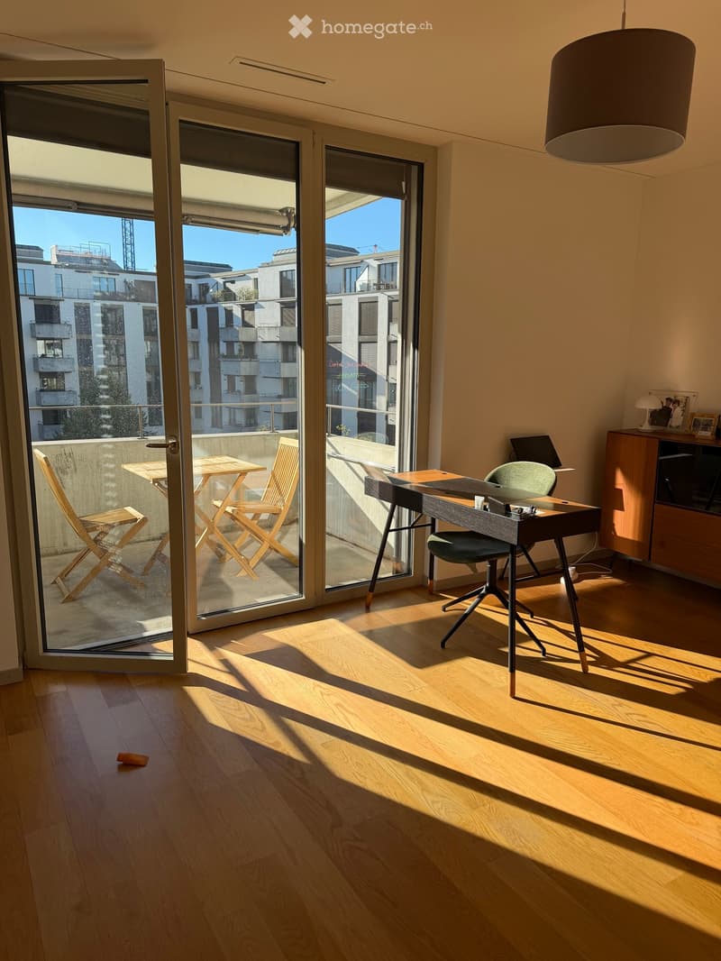 Bright room with wooden flooring, large sliding glass doors leading to a balcony, a desk with a chair, and a small table with chairs on the balcony.
