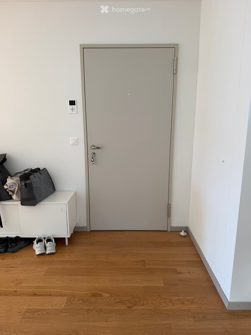 A small entryway with a wooden floor, white walls, a closed door, a small white bench holding bags and shoes, and a pair of white sneakers placed on the floor.