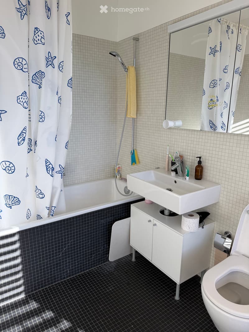 Modern bathroom with a bathtub, sink, toilet, and mirror. Features a white sink cabinet, a shower curtain with blue sea motifs, and a dark tiled floor.