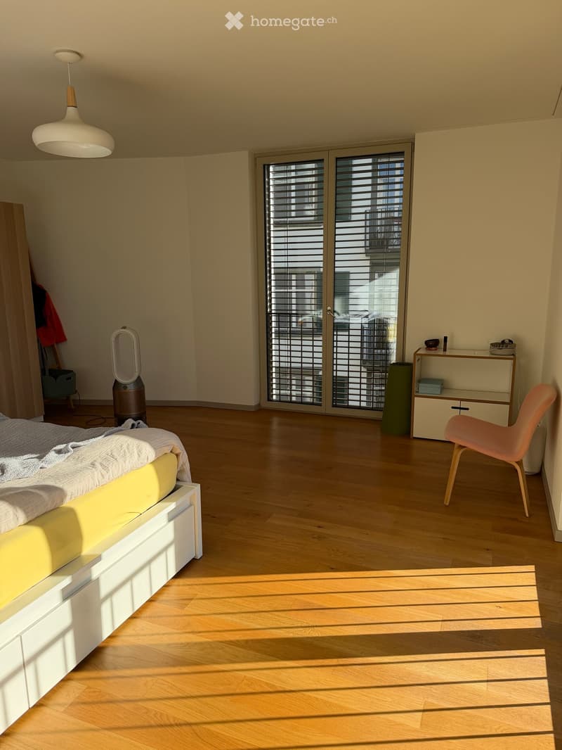 Modern bedroom with wooden flooring, a white bed with yellow headboard, a hanging lamp, a small table with a chair, and a fan near the window.