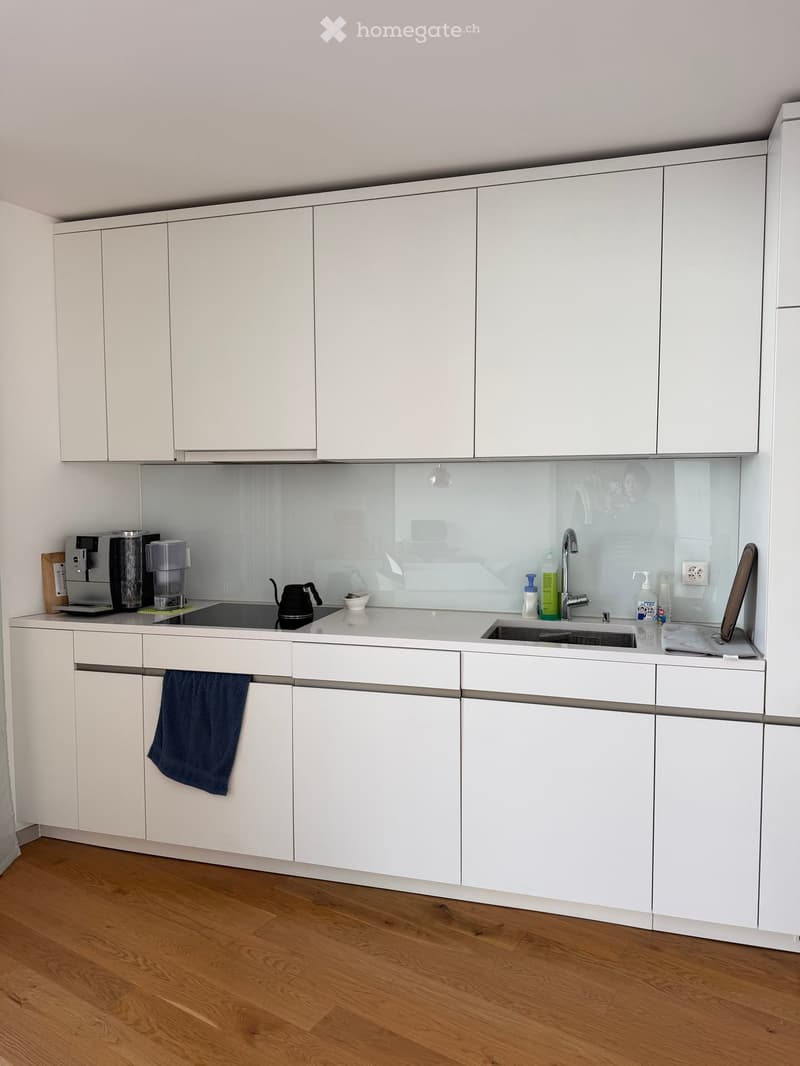Modern kitchen with white cabinets, wooden flooring, and a sleek countertop. Visible items include a coffee maker, teapot, sink, and various small appliances.