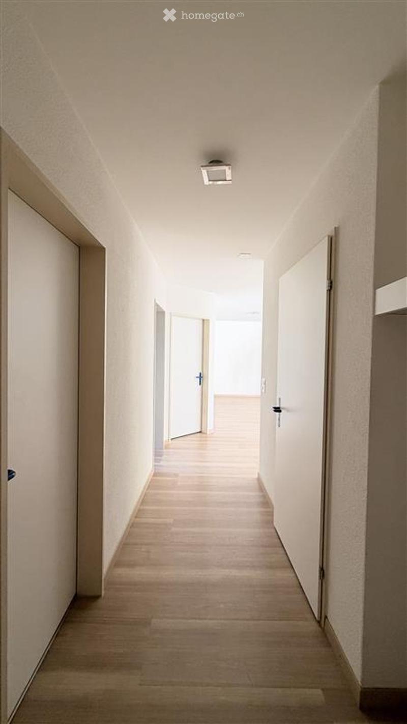 A clean, modern hallway with light wooden flooring, white walls, and recessed ceiling lighting. Doors are visible on both sides, leading to other rooms.