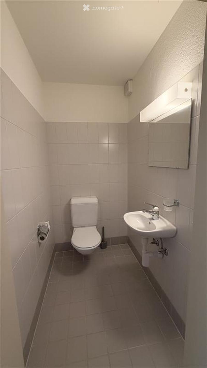 A compact bathroom with a toilet, sink, and mirror. The walls and floor are tiled in neutral tones, and the space is well-lit with a built-in light fixture above the mirror.